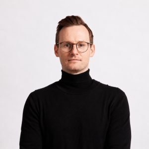 A professional portrait of a man wearing glasses and a black turtleneck, set against a plain white b.
